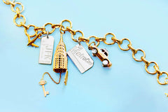 Gold charm bracelet with multiple NYC charms: silver JFK luggage tag, gold Chrysler Building with 'NYC' key, silver MetroCard, and gold taxi cab with silver taxi topper and silver wheels.