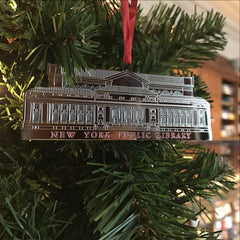 A metal holiday ornament in the shape of the New York Public Library building, with a silver finish, hanging on a Christmas tree.