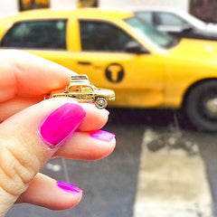 Close up photo of model's fingers holding gold vermeil taxi cab charm with silver taxi topper and silver wheels. Real taxi visible in background.