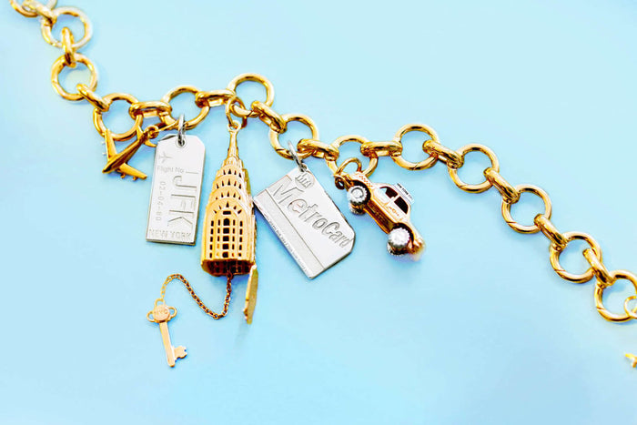 Gold charm bracelet with multiple NYC charms: silver JFK luggage tag, gold Chrysler Building, silver MetroCard, and gold taxi cab with silver taxi topper and silver wheels.